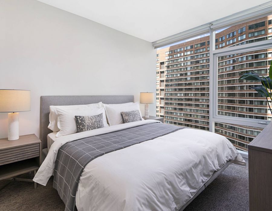Optima Signature in Downtown Chicago with Modern bedroom with a neatly made bed, two lamps, and large windows overlooking city buildings.